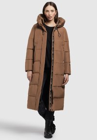 Brun puffjacka med huva, med en texturerad kviltad design, svarta detaljerade dragkedjor och en dragsko. Bärs över en svart tröja och mörka jeans.