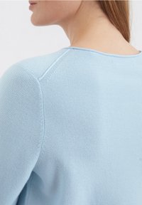 Hellblaue Strickjacke mit strukturiertem Finish, rundem Ausschnitt und Raglanärmeln. Das Material wirkt weich und atmungsaktiv.