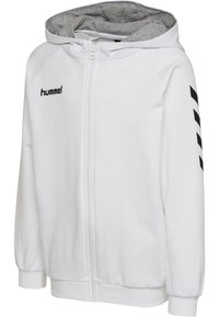 Haut à zip blanc fabriqué en matériau doux, avec une doublure de capuche grise, un logo noir sur la poitrine et des chevrons noirs le long des manches.