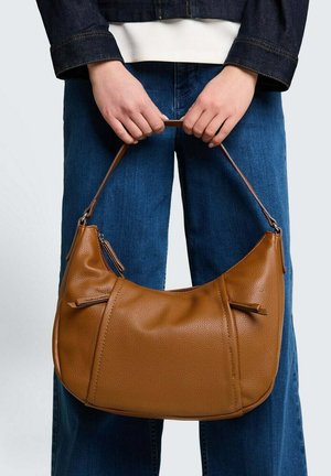 Person trägt blaue Jeans und dunkle Jacke und hält eine mittelbraune Lederhandtasche an ihrem kurzen Riemen auf Taillenhöhe.