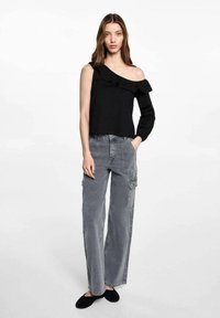 Crna off-shoulder majica s volanima, uparena sivoj širokim cargo hlačama s džepovima. Model nosi crne balerinke.