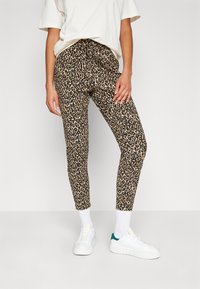 Leggings en imprimé léopard en tissu doux avec un design ajusté, arborant un motif brun et noir sur une base beige. Portés avec des baskets blanches.