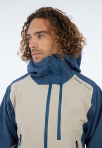 Lichte, bicolor jas in crème en marineblauw. Voorzien van een hoge kraag, trekkoorden en ritszakken aan de zijkant. Gestructureerde stof met ventilatie.