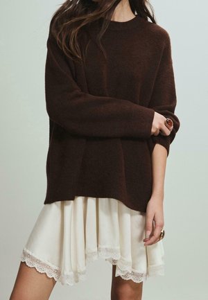 Kobieta w oversize'owym, ciemnobrązowym swetrze i kremowej spódnicy z koronkową lamówką, prezentująca pierścionki na obu dłoniach, stoi na neutralnym tle.