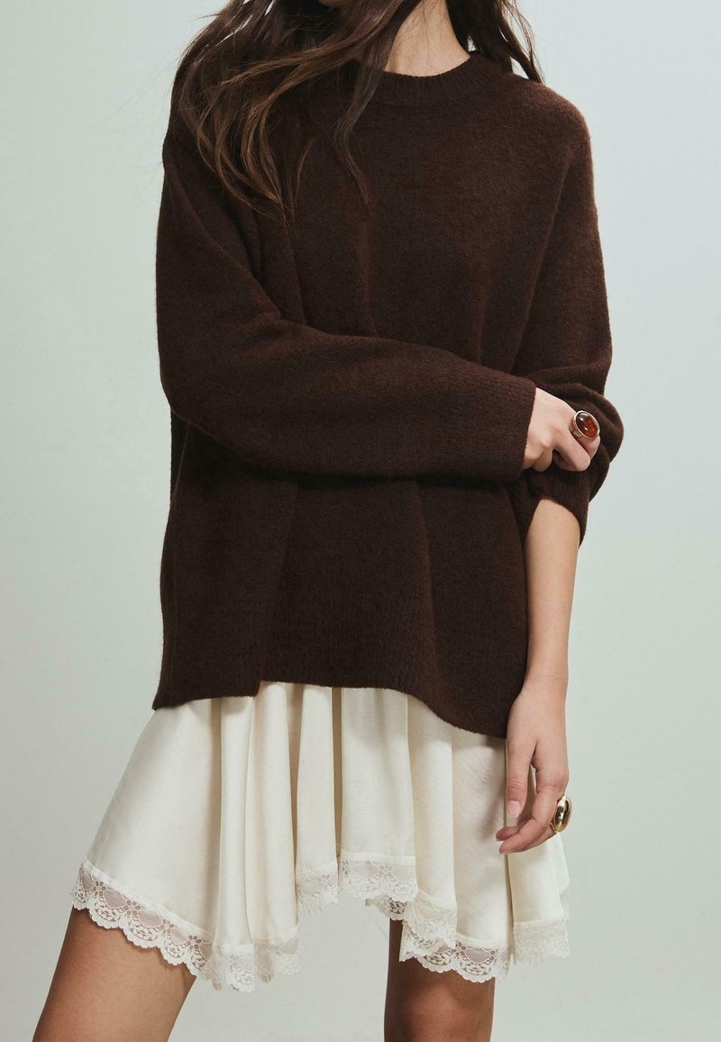 Femme portant un pull surdimensionné marron foncé et une jupe crème bordée de dentelle, montrant des bagues aux deux mains, debout devant un fond neutre.