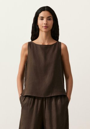 Žena s dlhými tmavými vlasmi, oblečená v bezrukávom hnedom topánke a zodpovedajúcich voľných nohaviciach, stojaca so založenými rukami v džepoch na jednofarebnom pozadí.