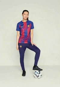 Frau in blau-rotem FC Barcelona-Trikot, die mit einem Fuß auf einem Fußball steht, vor einem schlichten hellen Hintergrund.