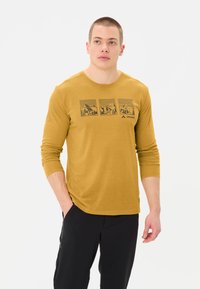 Chemise à manches longues moutarde avec un design graphique de montagnes et d'arbres, assortie à un pantalon noir, sur un fond neutre.