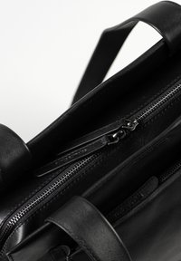 Close-up van een zwarte leren handtas met een dubbele ritssluiting en twee stevige handvatten op een witte achtergrond.