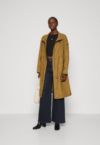 Cappotto lungo color marrone chiaro con cappuccio regolabile e tasche frontali, indossato su un top nero corto e jeans largo scuri con stivali neri con tacco.