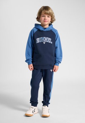 Barn med krøllet blondt hår iført en blå og marineblå Hummel hættetrøje, matchende bukser og hvide sneakers, stående mod en grå baggrund.