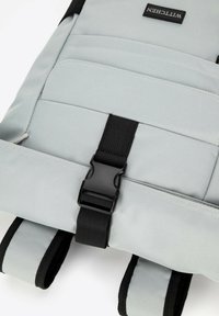 Sac à dos gris avec des accents noirs, doté d'une boucle centrale, de bretelles ajustables et de plusieurs poches avec des détails de couture visibles.