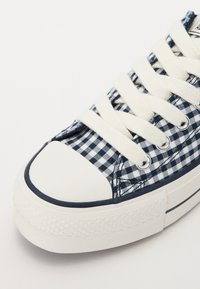 Zilā un baltā šahāre ar rakstu sporta apaviem ar baltu gumijas purngalu, kontrastējošu tumši zilu apdari un baltām šņorēm. Izteikta teksturēta gumijas zole.