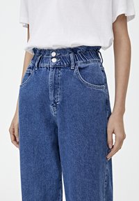 Jeans de mezclilla azul de talle alto con cintura elástica, dos botones, bolsillos delanteros y un corte relajado. La textura es ligeramente áspera.