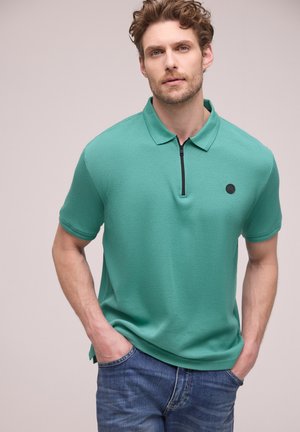 Mężczyzna o kręconych włosach, ubrany w miętową koszulkę polo z krótkim rękawem i czarnym zamkiem błyskawicznym oraz niebieskie dżinsy, stoi z rękami w kieszeniach na jasnym tle.