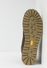 Ruskean vaelluskengän pohja syvien, karheiden urien kanssa; Vibram-logo keltaisena; rakenteellinen kumi pitävyyttä varten; vaalea ruskea väri.