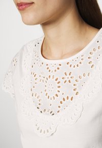Biały bawełniany top z misternym haftem kwiatowym i wycięciami wokół dekoltu i ramion. Gładka faktura i okrągły dekolt.