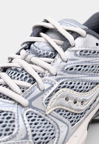 Scarpa atletica grigia con sezioni in rete, spesse lacci bianchi e una finitura lucida e testurizzata. Presenta un logo con un design a onda sul lato.