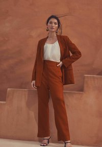 Veste de costume taillée de couleur rouille et pantalon taille haute, associés à un haut blanc et des sandales marines, devant un mur orange texturé.