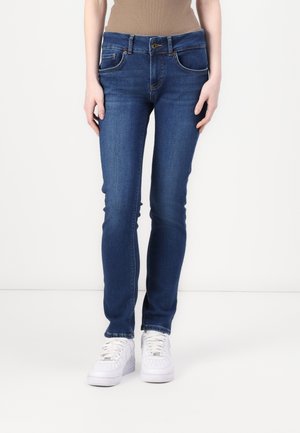 Jean skinny en denim bleu avec cinq poches, passants de ceinture et fermeture à bouton sur le devant. Porté avec des baskets blanches, montrant une texture lisse.