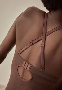 Gros plan du dos d'une personne portant un vêtement marron avec des bretelles croisées réglables et une fermeture à agrafe et œillet sur une découpe.