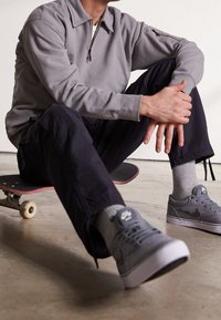Sudadera gris con diseño de medio cierre, combinada con pantalones cargo oscuros y zapatillas grises. Una patineta yace debajo de la figura sentada.