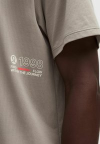 Beżowa, teksturowana koszula z krótkim rękawem z napisem "1998 FLOW FIND WITHIN THE JOURNEY", noszona przez osobę o ciemnej karnacji.