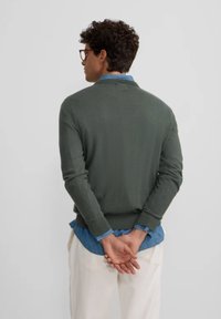 Suéter verde de punto con un cuello redondo, puños y dobladillo acanalados. Llevado sobre una camisa de chambray azul clara, visto desde la parte de atrás.