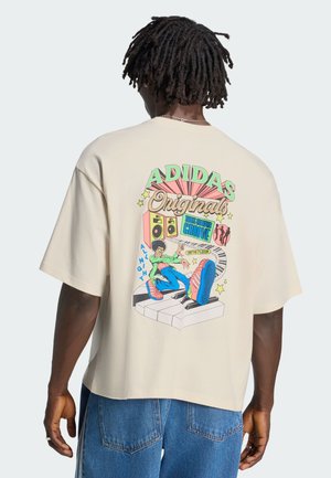 Mann mit Dreadlocks trägt ein beiges Adidas Originals T-Shirt mit buntem Retro-Tanz- und Musikmotiv auf dem Rücken und blaue Jeans.