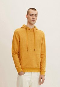 Sudadera amarilla de punto con un patrón texturizado, capucha con cordón, bolsillo frontal, y puños y dobladillo acanalados. Combinada con pantalones blancos.