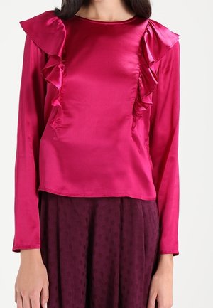Kvinde iført en skinnende magenta bluse med lange ærmer og flæser foran, kombineret med en mørkelilla tekstureret nederdel.
