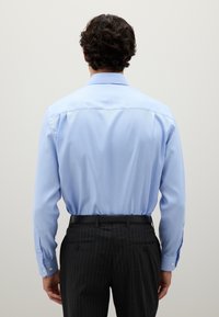 Uomo con capelli ricci e scuri che indossa una camicia azzurra chiara infilata in pantaloni scuri a righe grigie con una cintura nera, girato di spalle.