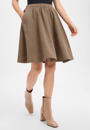 Femme portant une jupe à carreaux marron et noire jusqu'aux genoux et des bottines beiges à talons, debout sur un fond blanc.