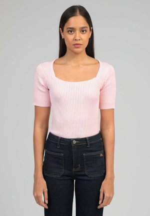 Jeune femme aux cheveux raides et foncés portant un haut rose clair côtelé à manches courtes et un jean bleu foncé, debout devant un fond gris uni.