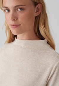 Beige knit sweater with a high collar, featuring a soft texture and subtle variations in color. Close-up of face with light skin and blonde hair.