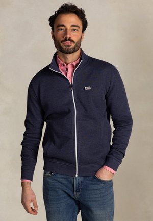 Man draagt een marineblauw jack met rits over een roze overhemd en een blauwe spijkerbroek, staat met één hand in de zak tegen een effen achtergrond.