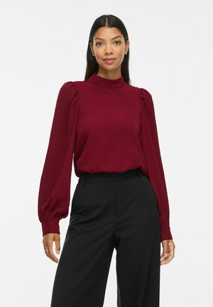 Vrouw met lang donker haar, gekleed in een bordeauxrode blouse met lange mouwen en opgepofte schouders en zwarte hooggetailleerde broek, staand tegen een effen achtergrond.