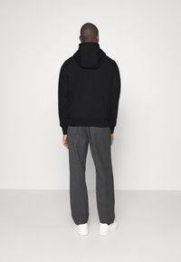 Uomo che indossa un felpa nera con cappuccio, pantaloni grigi e sneakers bianche, in piedi di fronte a un muro grigio chiaro uniforme.