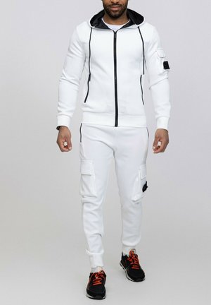 Haut à zip blanc avec des bordures et des cordons noirs, assorti à un jogger blanc avec des poches latérales. Baskets noires et orange.