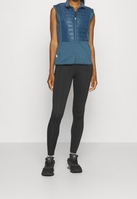 Ärmlös väst i mörkt teal med kviltad front, dragkedja och sidofickor, kombinerad med svarta leggings och svarta träningsskor.