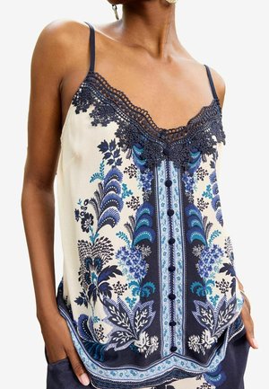 Vrouw die een mouwloze crèmekleurige top draagt met een marineblauw bloemenpatroon, kanten rand langs de halslijn en knoopdetails aan de voorkant.