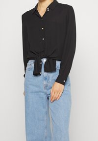 Donna che indossa una camicia nera a maniche lunghe con bottoni legata in vita, abbinata a jeans blu chiaro a vita alta.