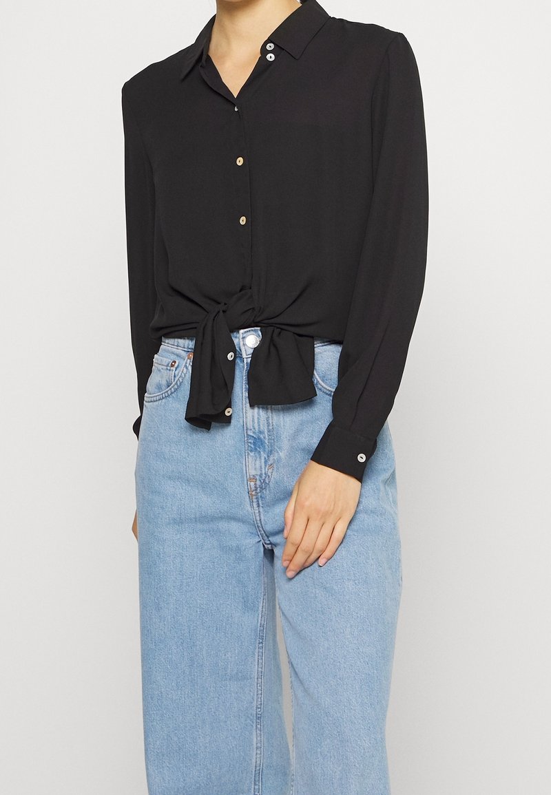 Donna che indossa una camicia nera a maniche lunghe con bottoni legata in vita, abbinata a jeans blu chiaro a vita alta.