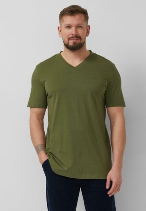 Mężczyzna z brodą w oliwkowozielonej koszulce z krótkim rękawem i dekoltem w serek oraz w ciemnych spodniach, stojący z jedną ręką w kieszeni na szarym tle.