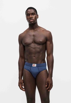 Homme musclé debout, faisant face, portant un slip bleu avec une étiquette blanche sur la ceinture, sur un fond blanc uni.