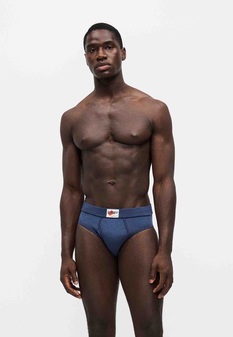 Homme musclé debout, faisant face, portant un slip bleu avec une étiquette blanche sur la ceinture, sur un fond blanc uni.