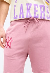 Άτομο που φορεί μπεζ μπλούζα Los Angeles Lakers και ροζ σορτς με κόκκινο λογότυπο New York Yankees και κορδονάκι στη μέση.