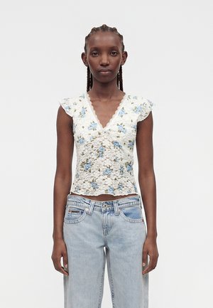 Woman with braided hair wearing a white lace floral top with blue flowers and light blue denim jeans, standing against a plain background.