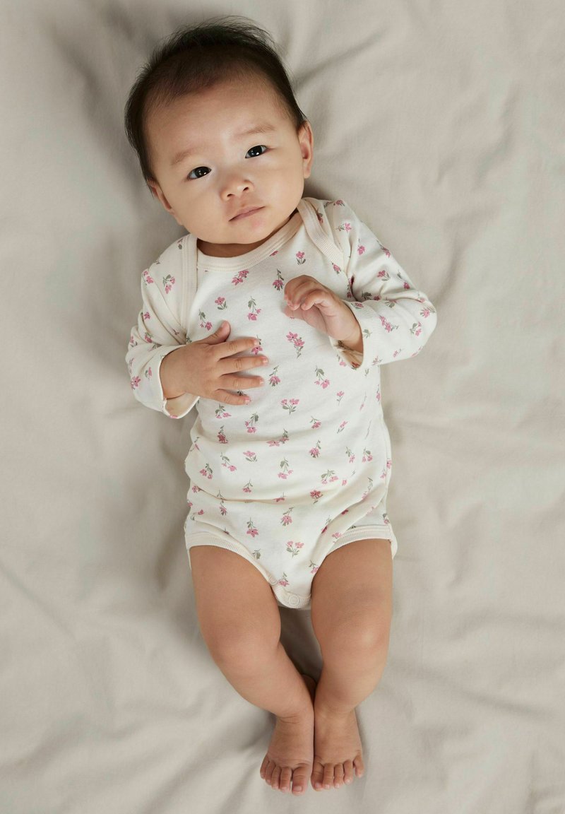 Lyse babyheldragt med lille pink blomstermønster, lange ærmer og tryklås. Blødt stof mod en beige baggrund.