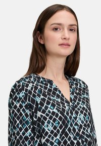 Blouse à manches longues en noir avec un motif géométrique blanc et sarcelle. Présente un décolleté en V et une texture de tissu lisse.